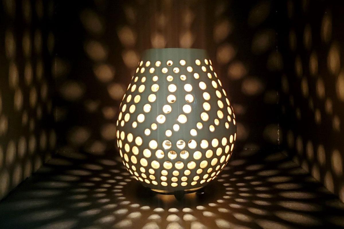 A pottery piece with holes making a spiral design with a light shining through it against a wall